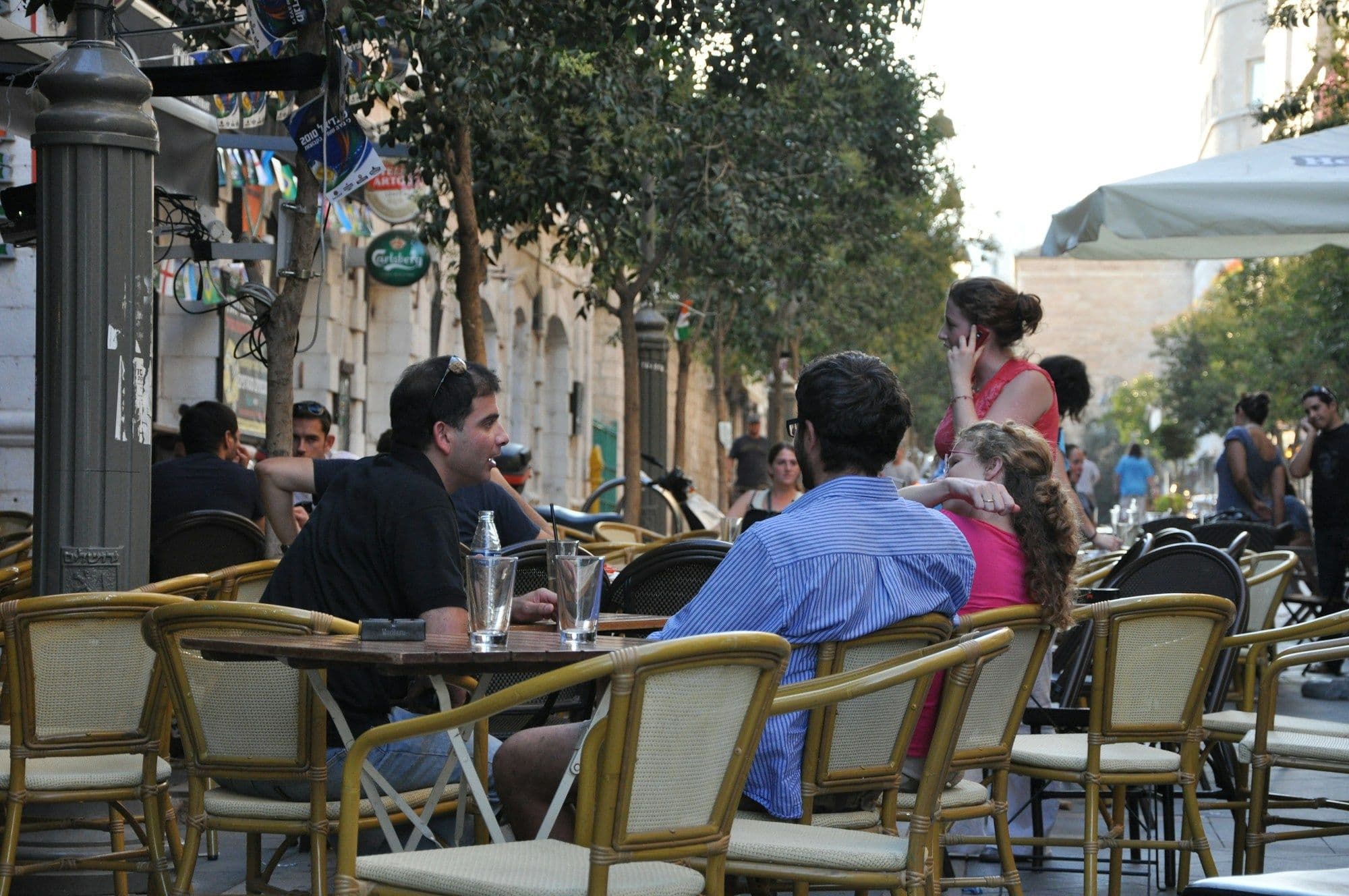 People talking over drinks at wicker-chair tables on a tree-lined pavement café terrace in the late afternoon.