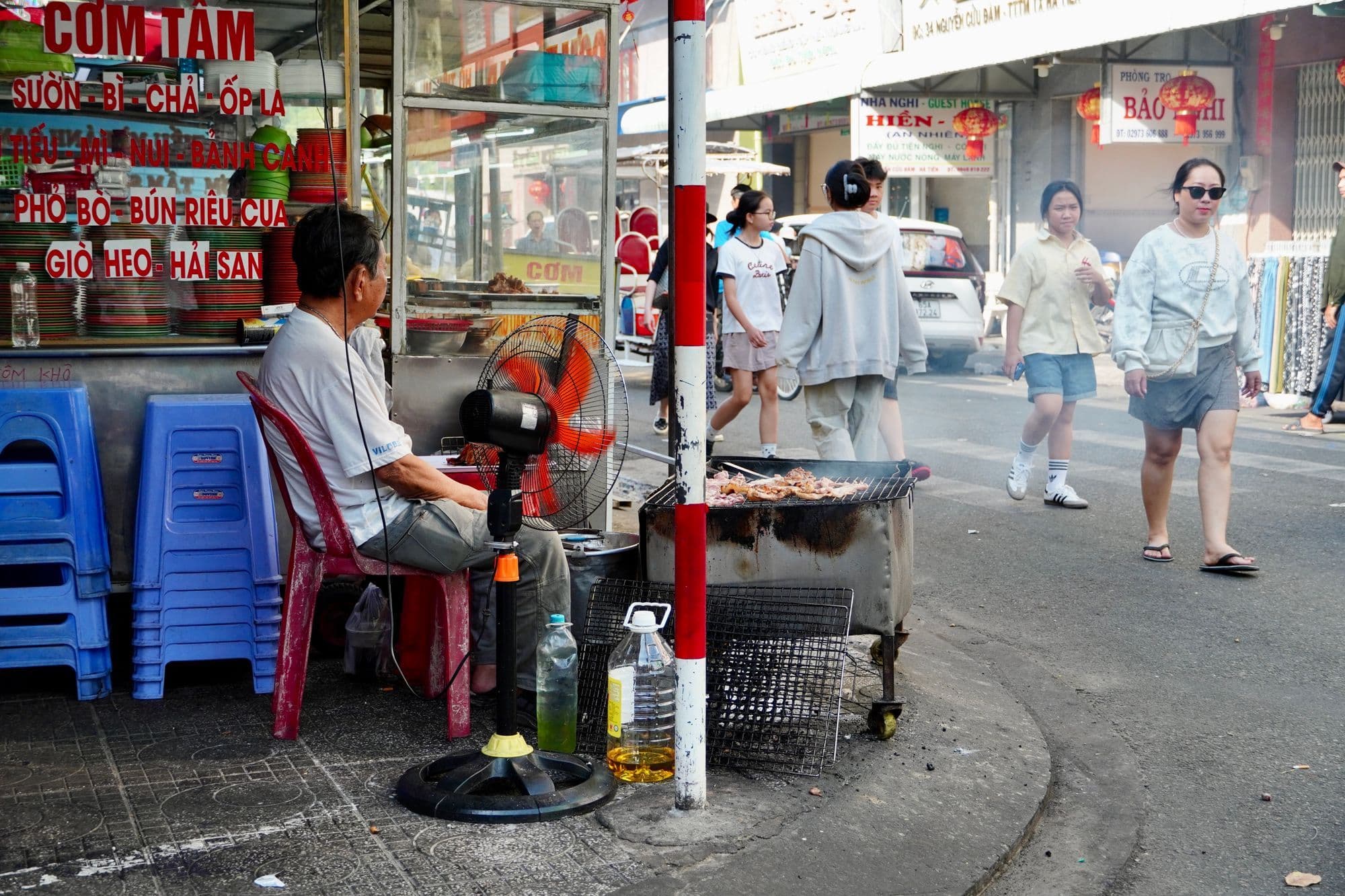 A street vendor grills meat on a small charcoal stove beside a cơm tấm stall with blue plastic stools, as people walk past.