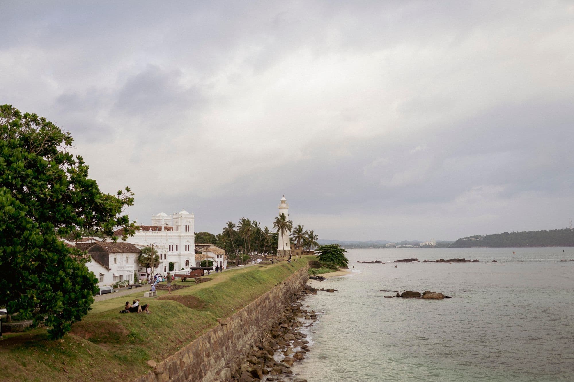 The grassy rampart of Galle Fort running along the sea towards a white lighthouse and colonial buildings under a cloudy sky.