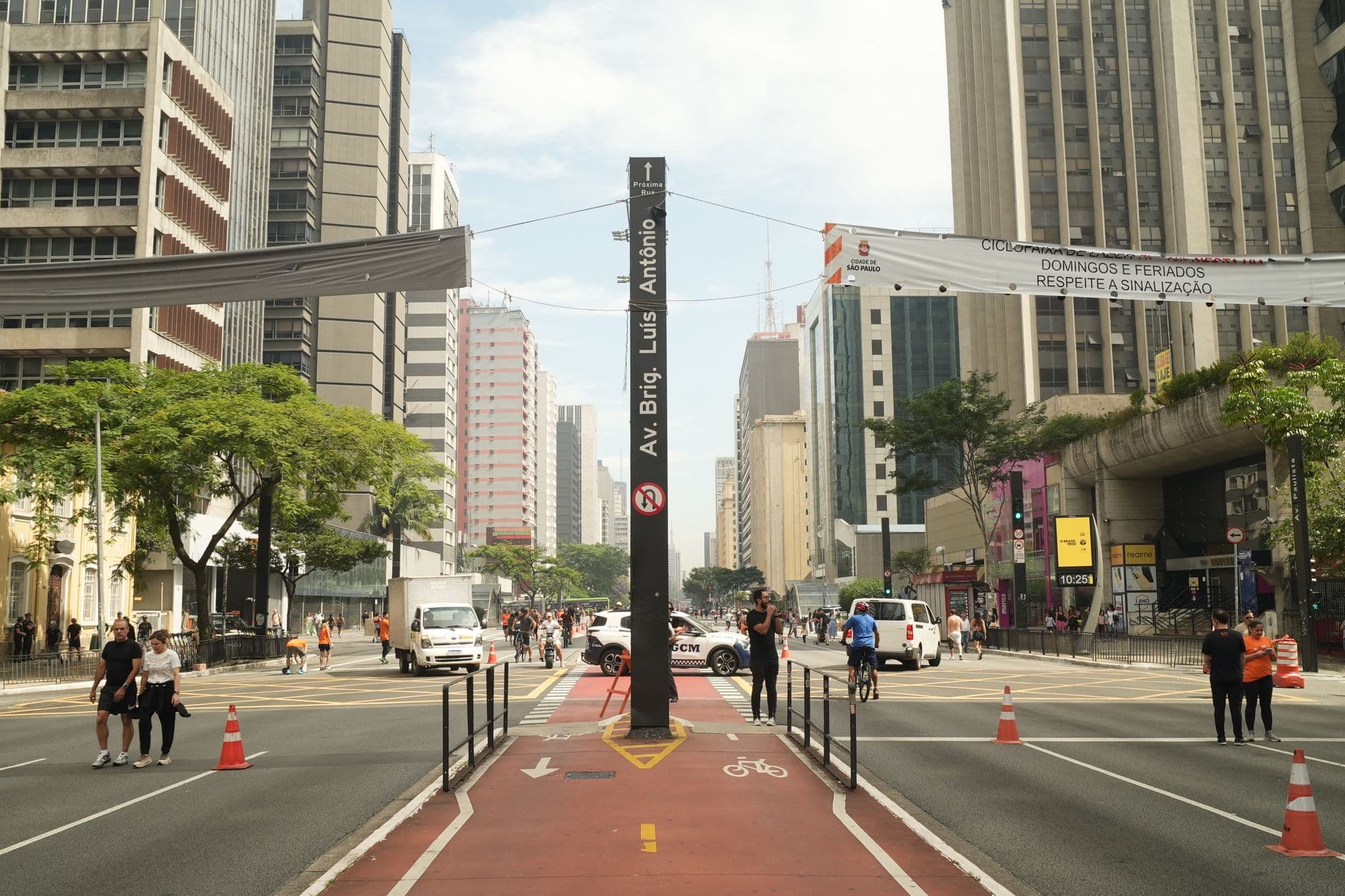 Pedestrians and traffic moving between tall buildings on Avenida Paulista in daylight.