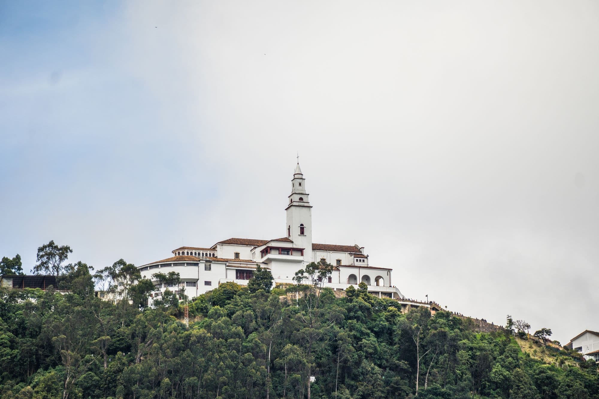 The white church and buildings of the Monserrate sanctuary on top of a forested hill above Bogotá under a pale sky.
