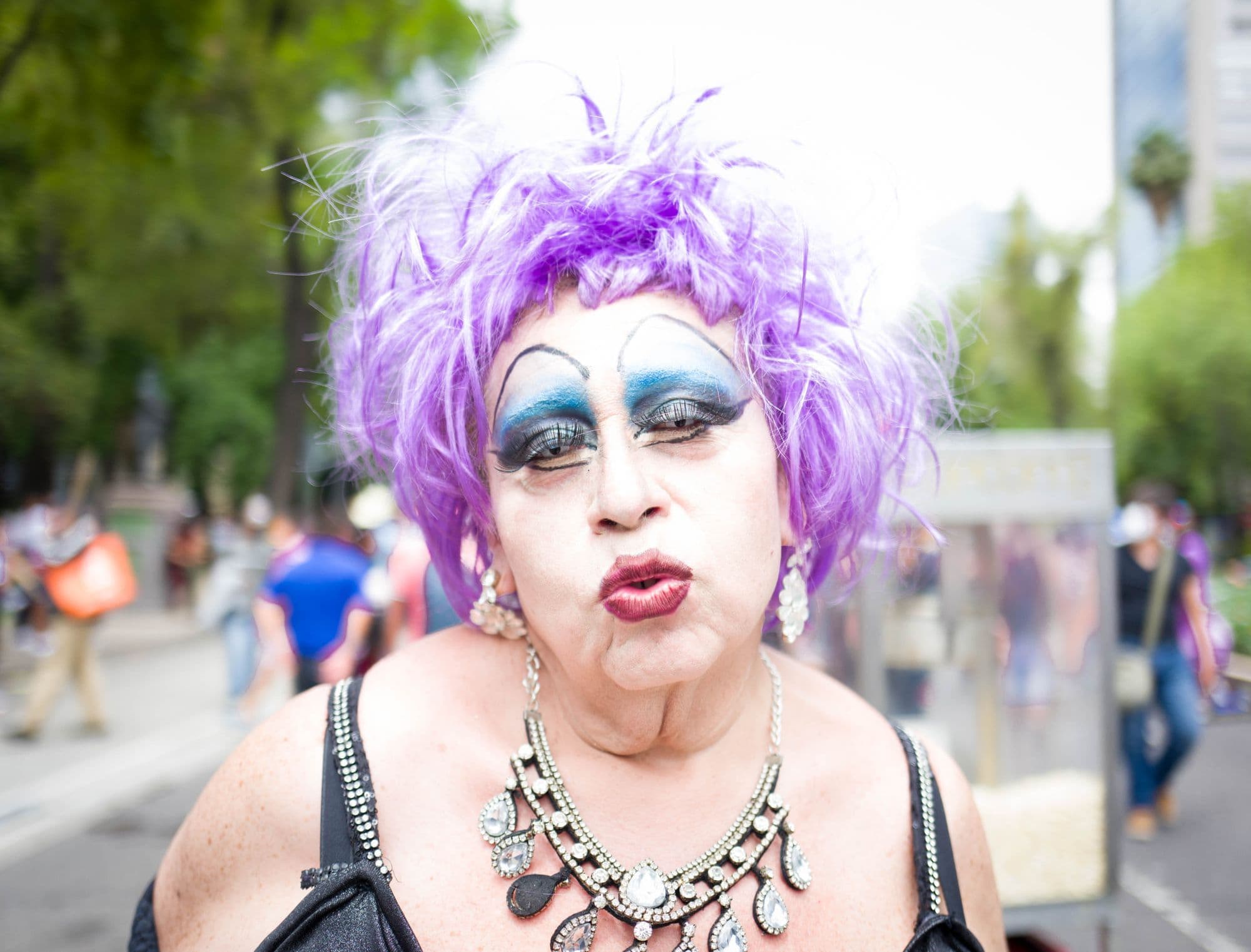 A drag performer with a purple wig, blue eyeshadow and a crystal necklace on a tree-lined avenue during Pride.