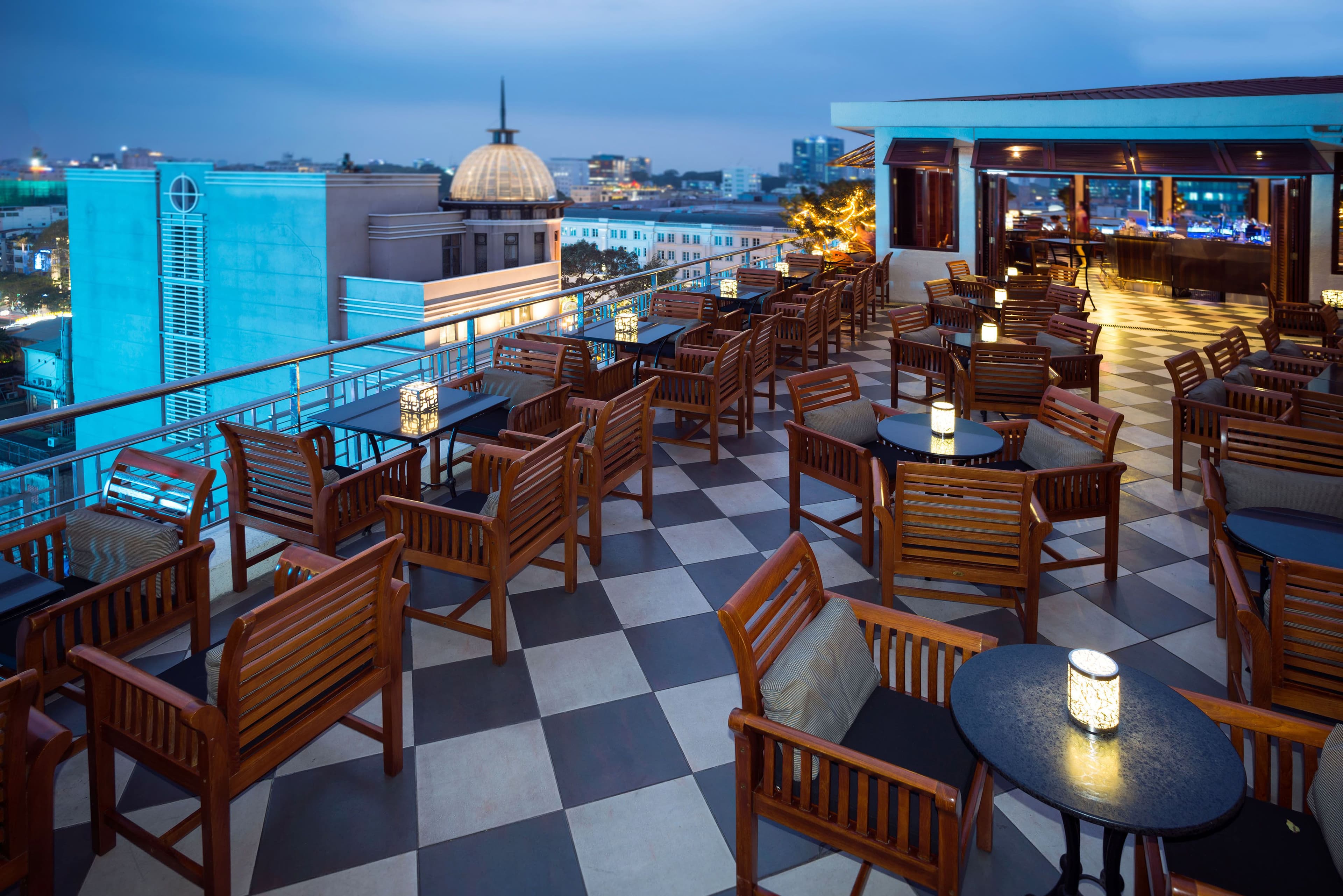 Wooden armchairs and candlelit tables on a checkerboard rooftop terrace at dusk, with a domed building beyond.