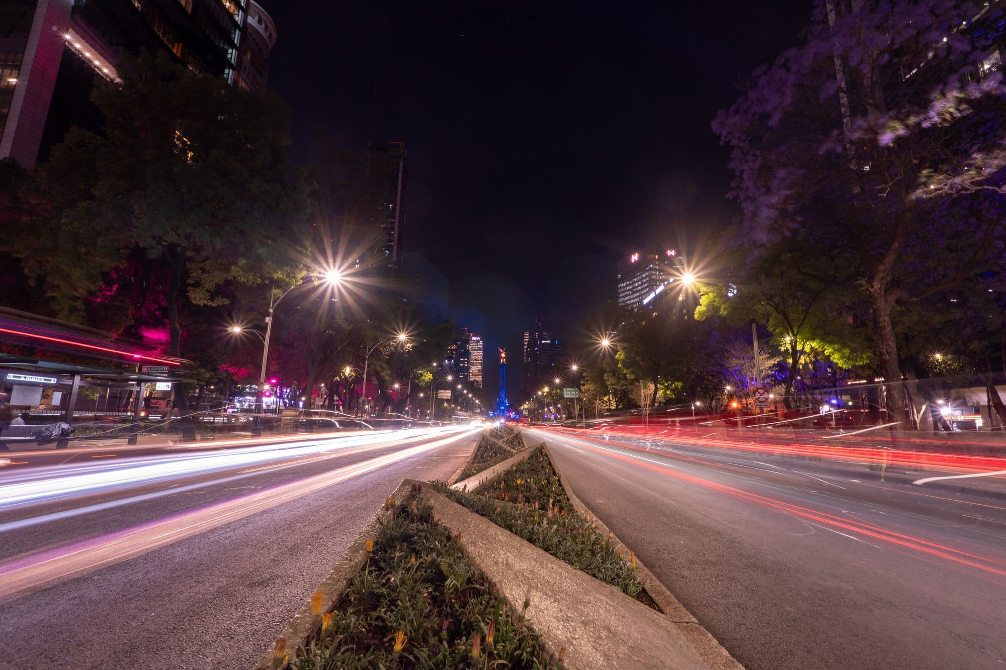 Long-exposure light trails of traffic on Paseo de la Reforma at night, with the Ángel lit blue in the distance.