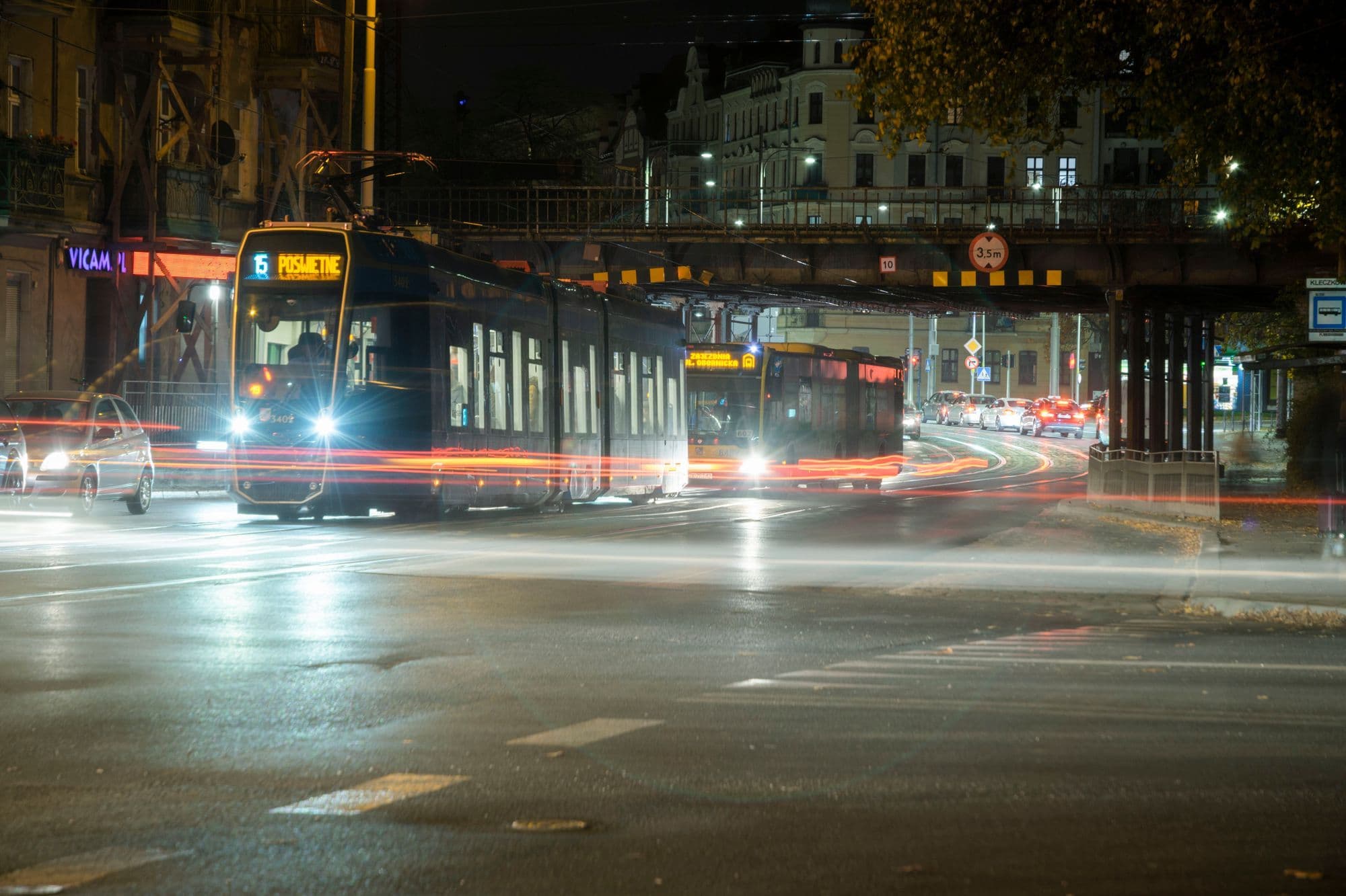 A tram passes through a city street at night, leaving trails of light.