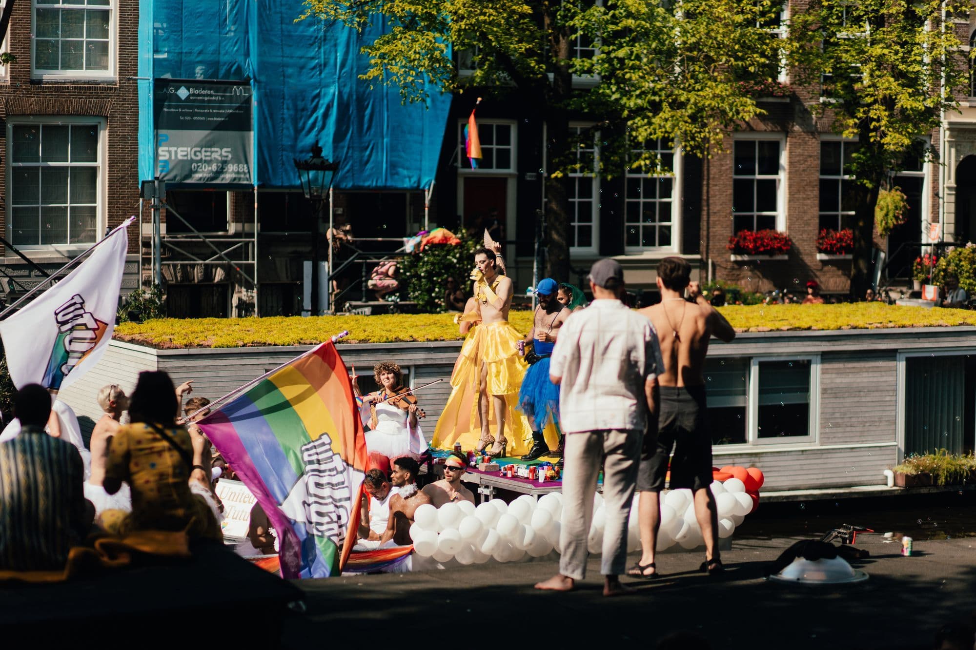 Decorated boats and a crowd celebrating along an Amsterdam canal during Pride.