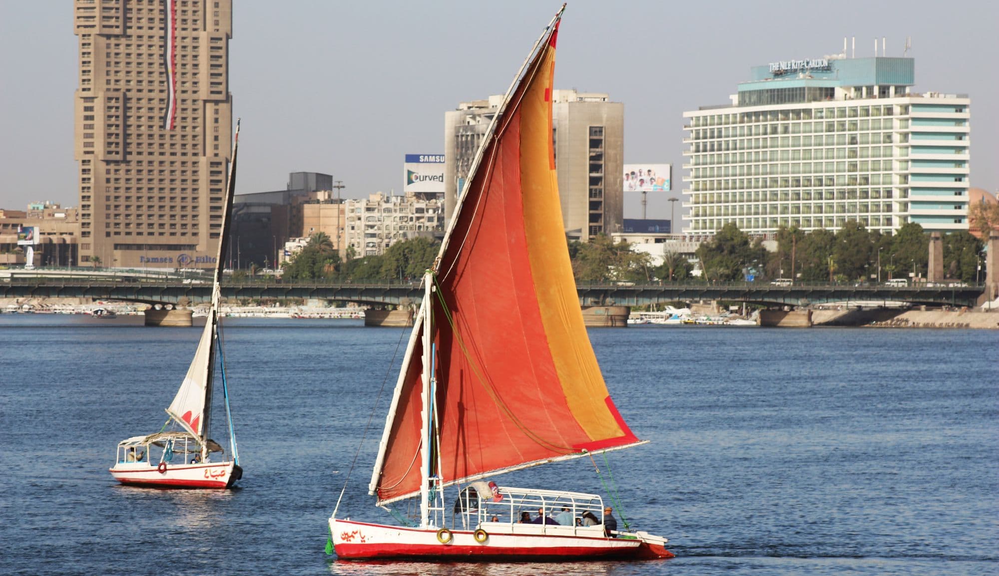 Two feluccas with orange and white sails on the Nile in Cairo, with a bridge and tall riverside hotels behind.