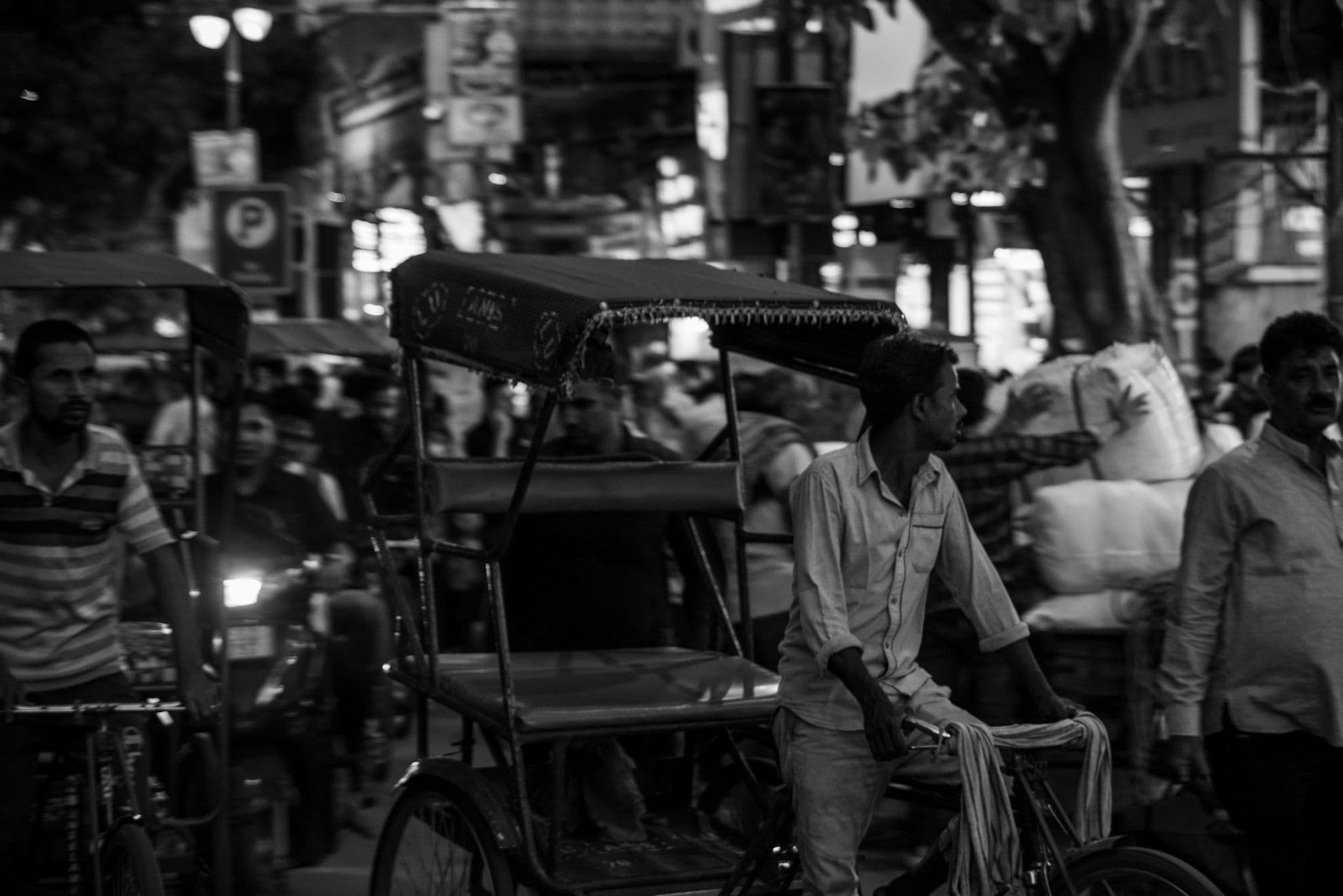 A black-and-white night photo of a cycle rickshaw driver waiting in a crowded Old Delhi street under hanging lights.