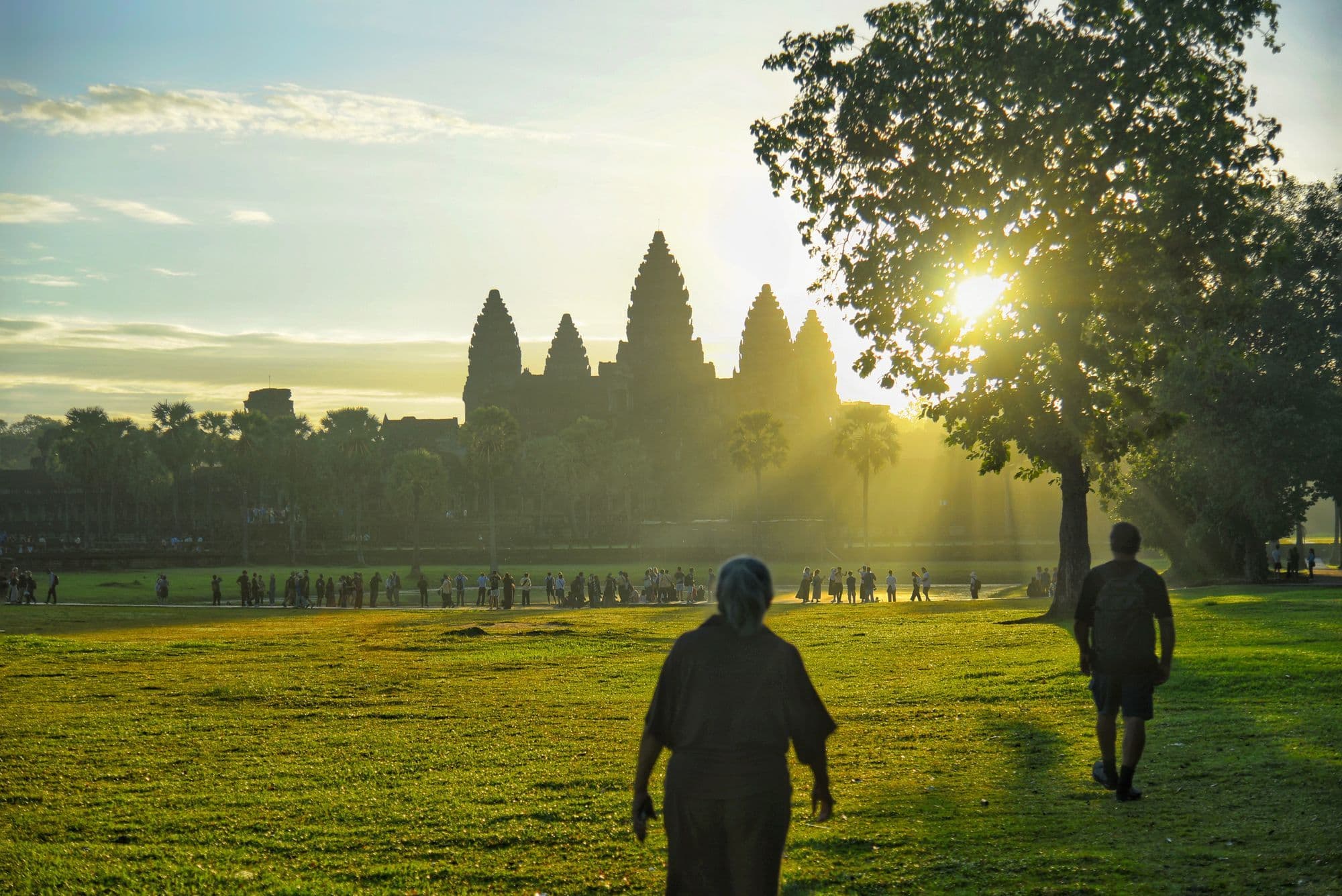 Silhouetted visitors walk along the causeway towards the dark outline of Angkor Wat under a bright sunrise sky.