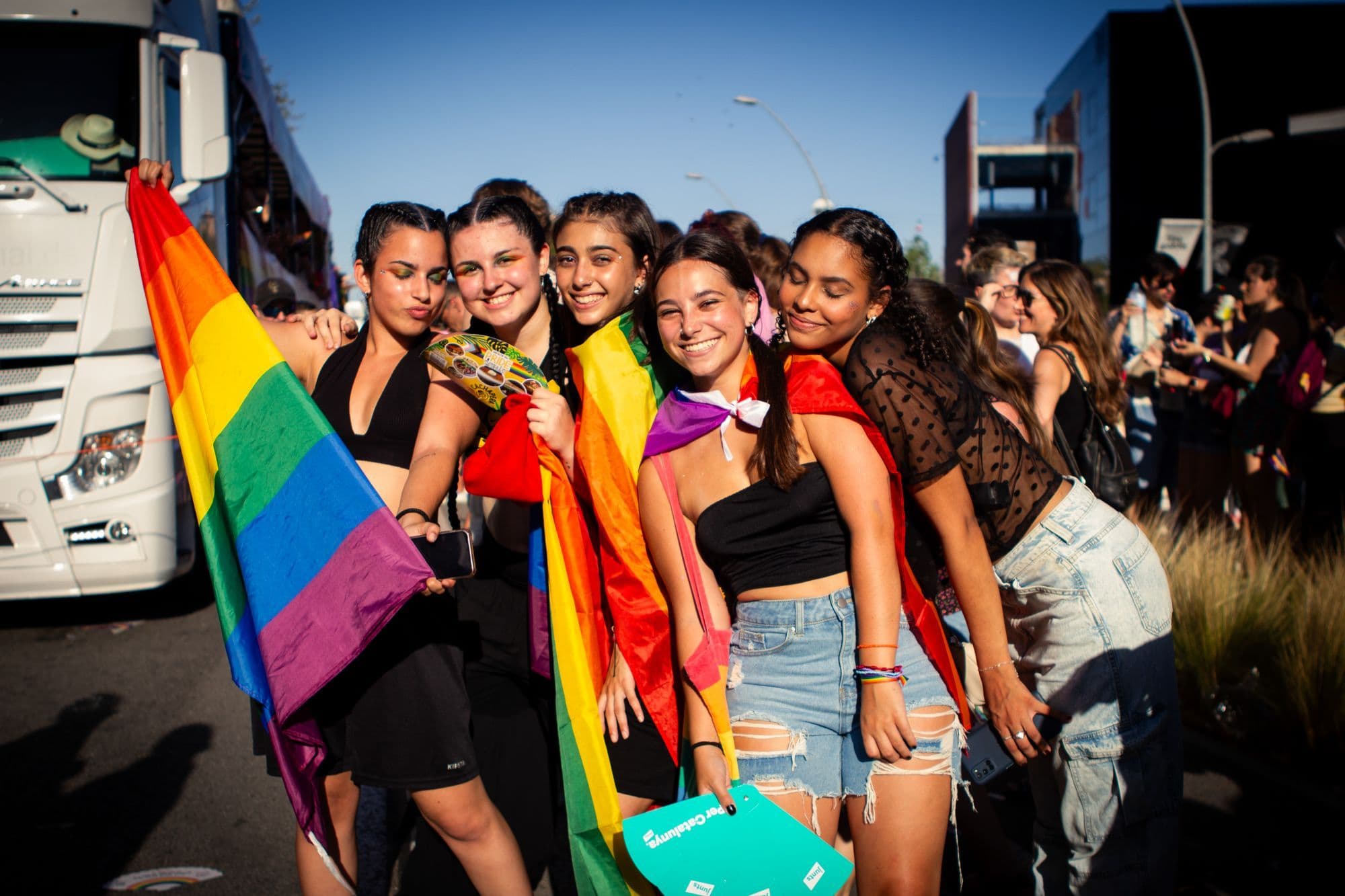 A group of young women wrapped in rainbow flags smiling together on a Barcelona street during Pride.